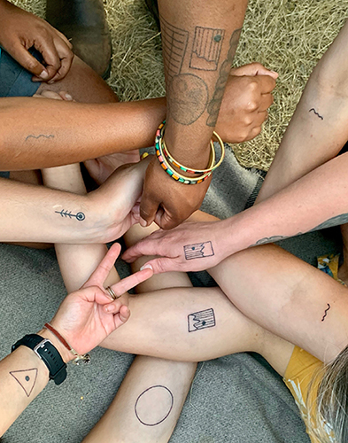 Arms and hands with many different skin tones come together. Some hands touch each other, or make bunny ears, or a fist. Each limb is adorned with ink medicine by Jaz Whitford